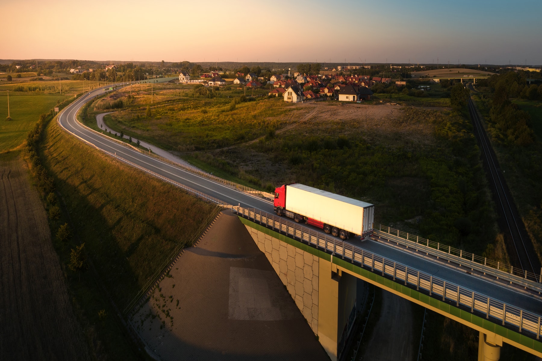 Hva er HVO100 - luftfoto av en lastebil som kjører på en landevei i solnedgangen, med en by og en bro i et nordisk landskap – et symbol på en vognpark som kjører på fornybar diesel