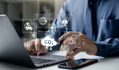 Scope 1 2 og 3-utslipp: En fagperson arbeider på en bærbar datamaskin, omgitt av digitale ikoner for CO2-utslipp, en fabrikk, fornybar energi og en elbil - et bilde på den digitale sporingen som ligger bak bedrifters utslippsrapportering