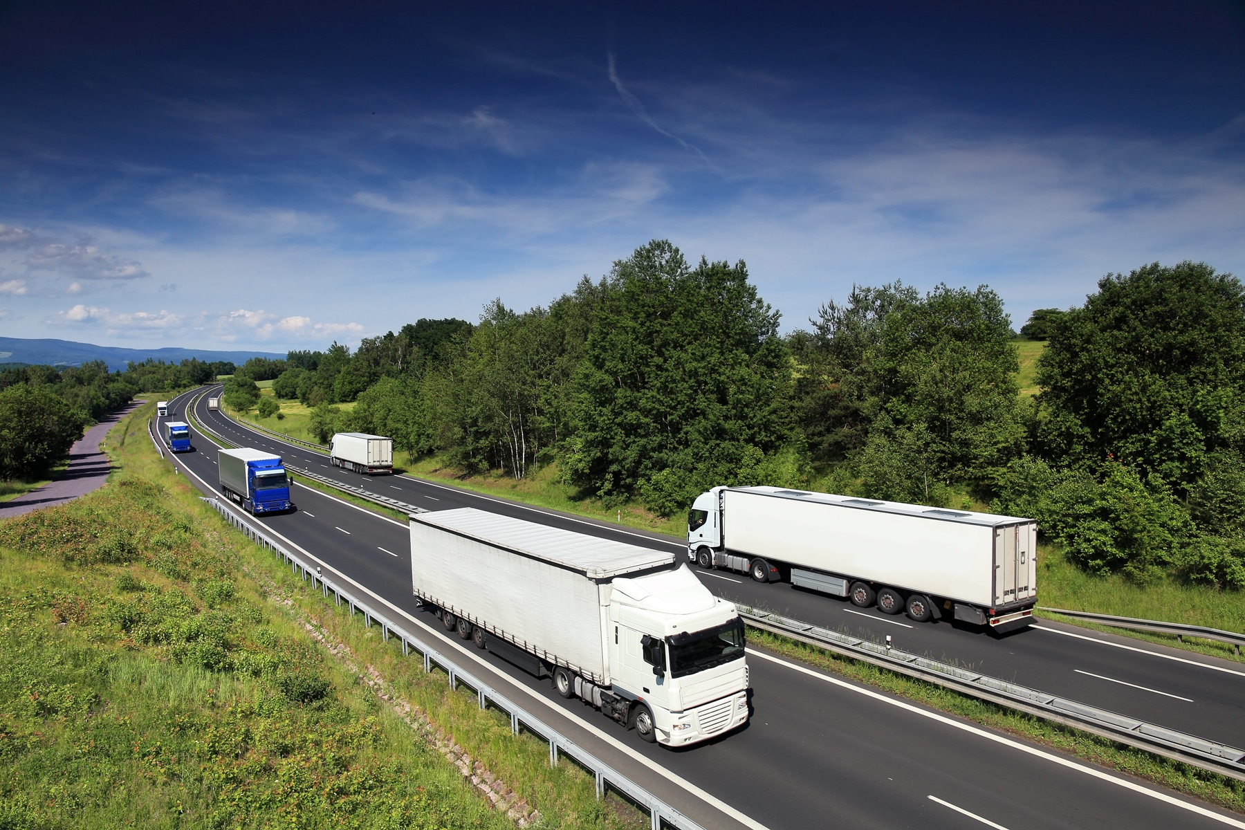 Klimaregler i transport - flere lastebiler kjører på en flerfelts motorvei gjennom grønt kupert landskap – et bilde på den veitransporten som omfattes av EUs klima- og utslippsregler